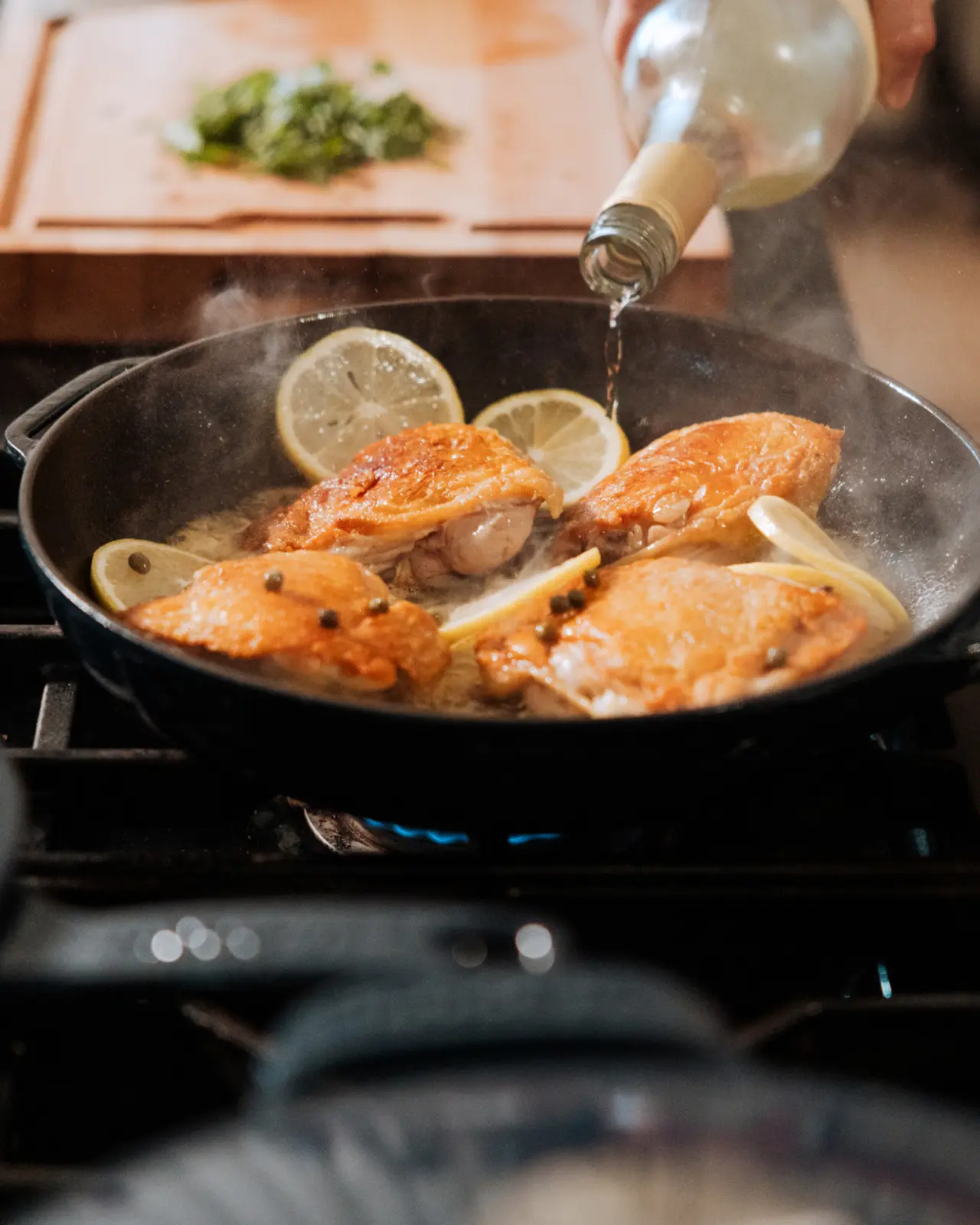 Juice is being poured over sizzling chicken breasts in a pan, accompanied by lemon slices and herbs in the background.