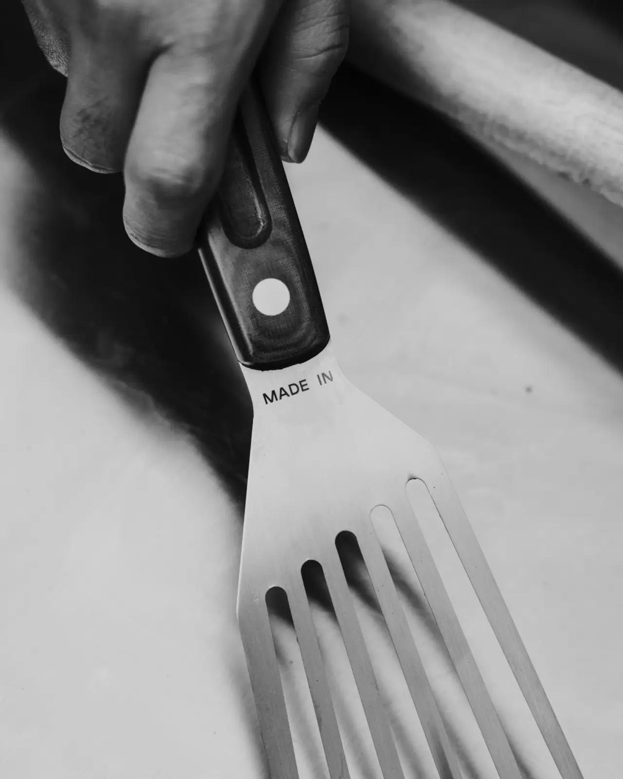 A person's hand grips a large stainless steel fork with "MADE IN" text imprinted on the handle, set against a blurred background.