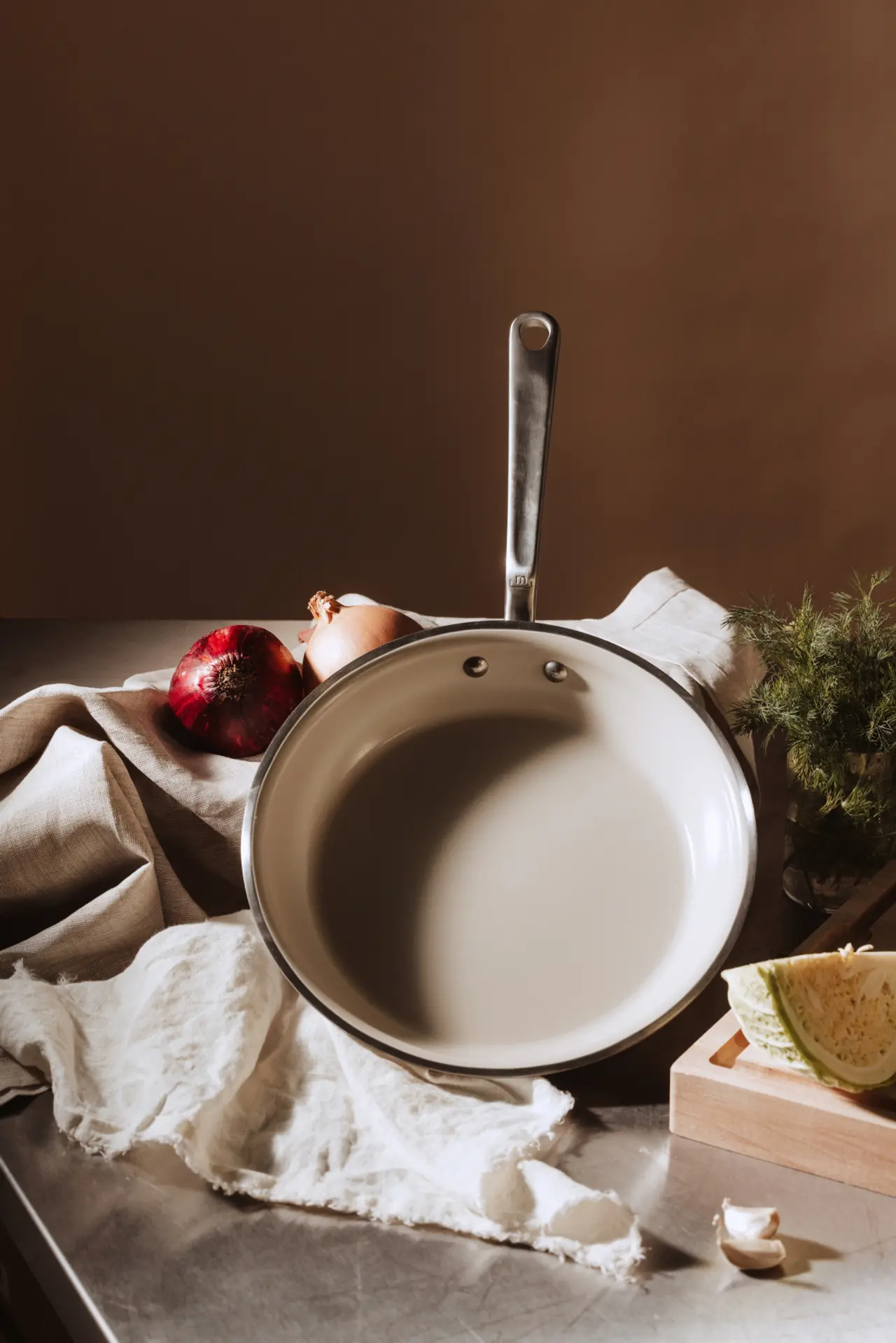 A stainless steel saucepan sits on a textured surface surrounded by fresh ingredients, including red onions, garlic, and herbs.