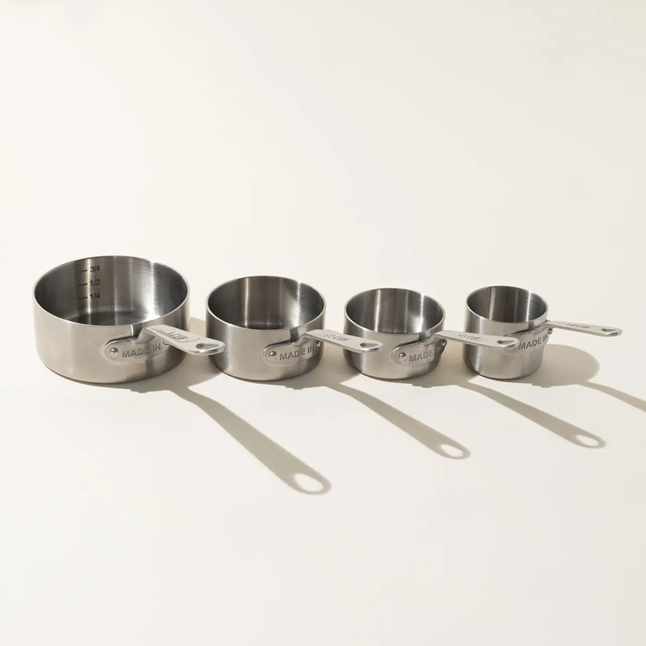 Four graduated stainless steel measuring cups are aligned in a row, decreasing in size from left to right, casting slight shadows on a bright surface.