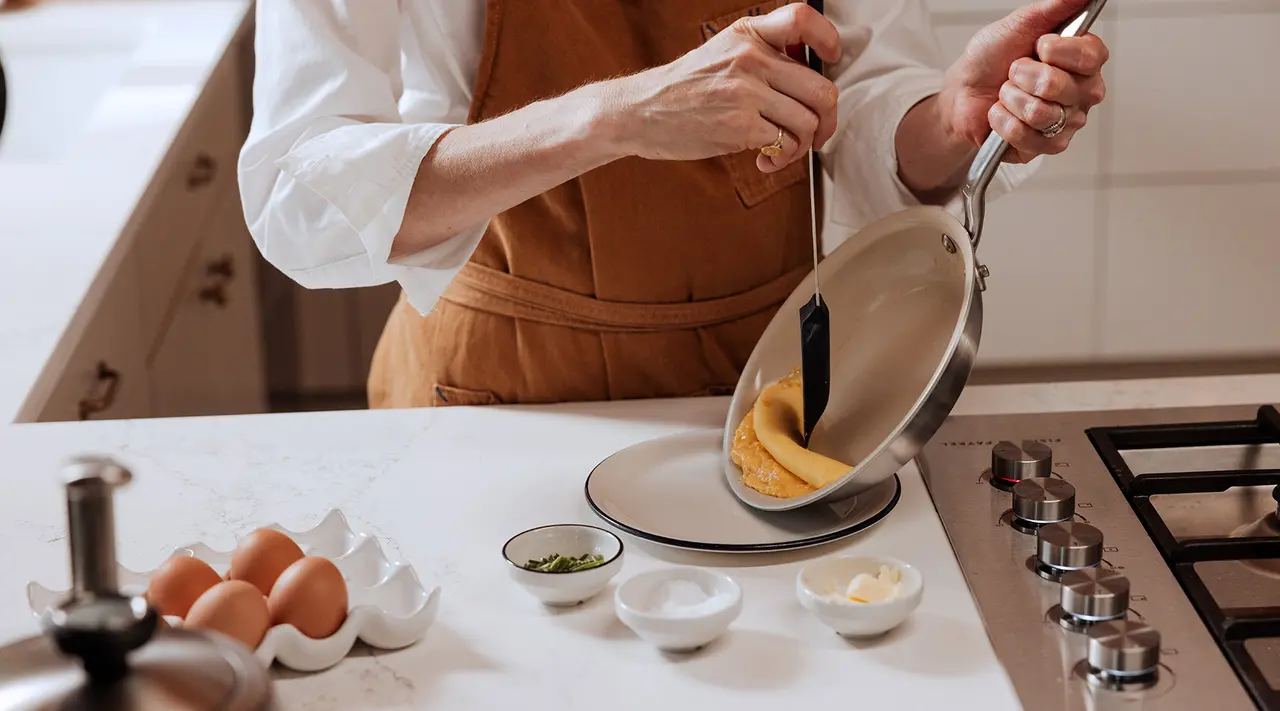 A person in an apron is pouring a pancake batter onto a plate from a small pan, with ingredients arrayed on the counter.