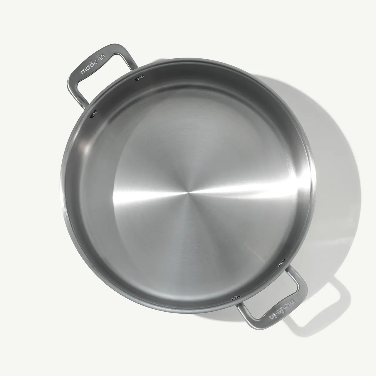 A stainless steel frying pan with two handles is shown from above on a light surface.