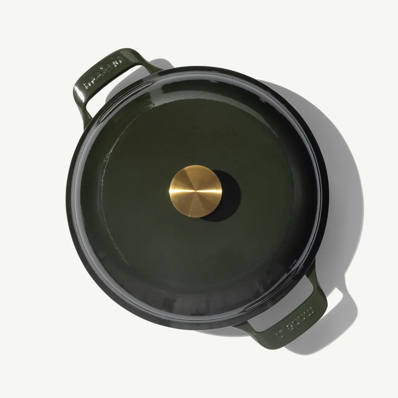 A round, dark green cooking pot with a brass knob on its lid sits on a light background.