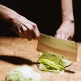 A hand using a large knife to chop a head of cabbage on a wooden cutting board.