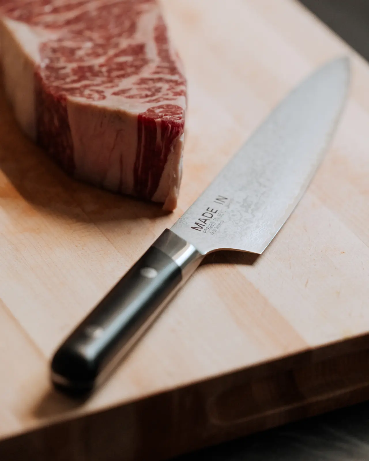 A chef's knife lies next to a block of uncooked steak on a wooden cutting board.