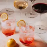 A selection of cocktails and glasses of wine is arranged on a wooden surface, featuring two drinks with ice and garnished with lemon slices.