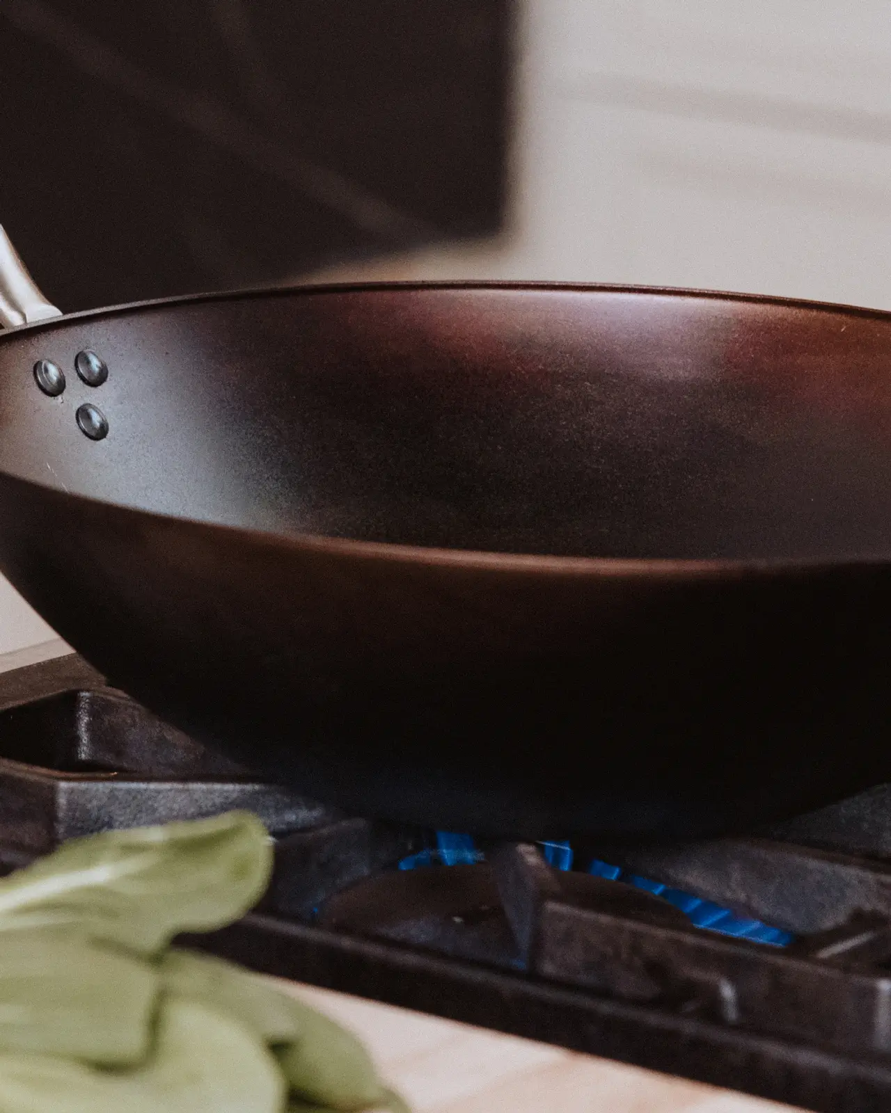 A black wok sits on a stove with blue flames visible beneath it, accompanied by green vegetables in the foreground.