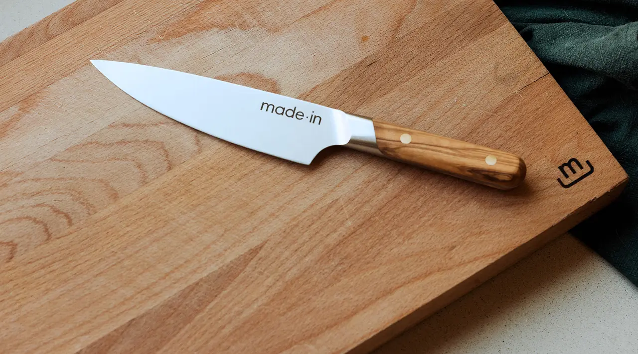 A chef's knife with a wooden handle labeled "made.in" is placed diagonally on a wooden cutting board on a countertop with a green cloth partially visible in the corner.
