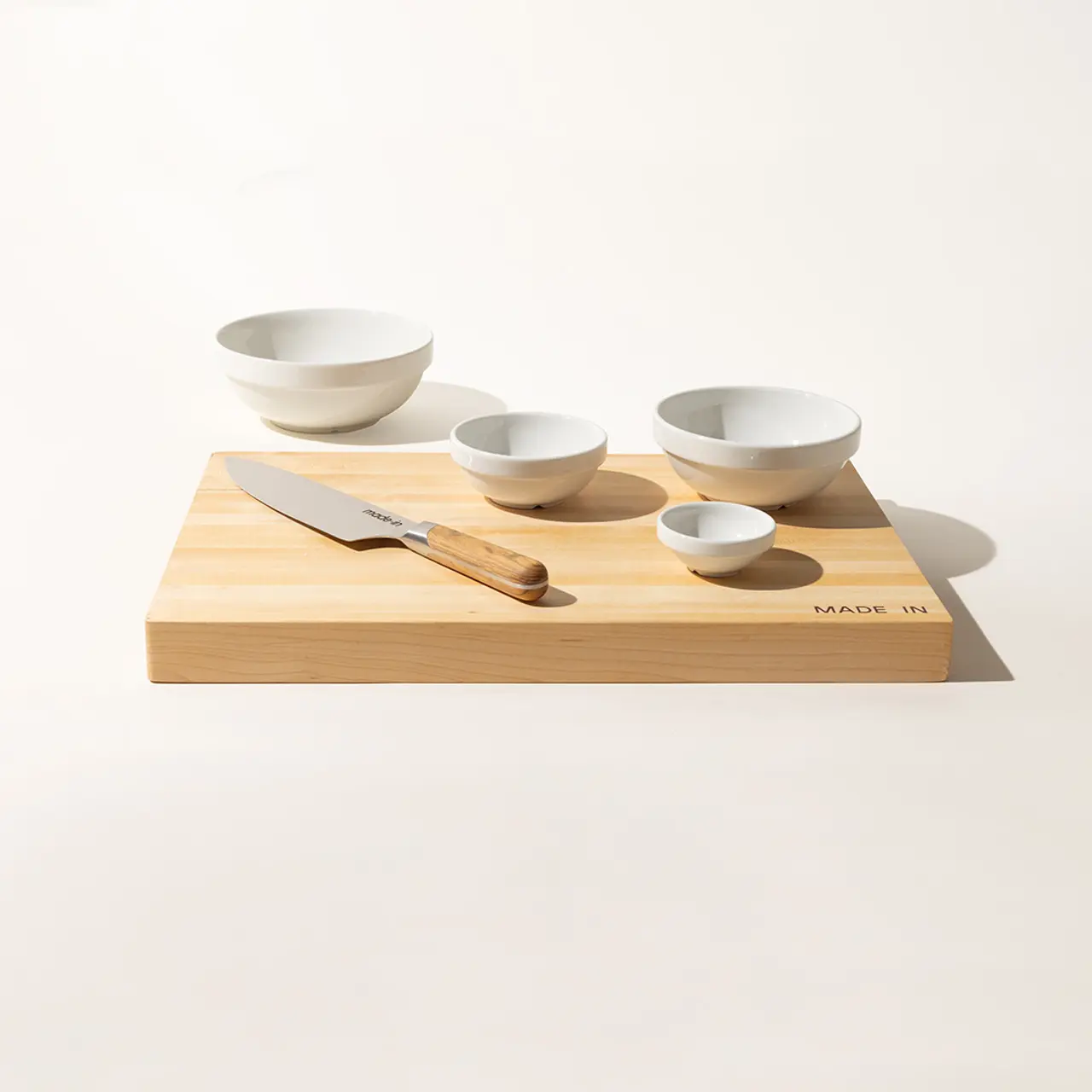 A neatly arranged set of ceramic bowls and a wooden spoon on a bamboo cutting board with a kitchen knife, all on a light background.