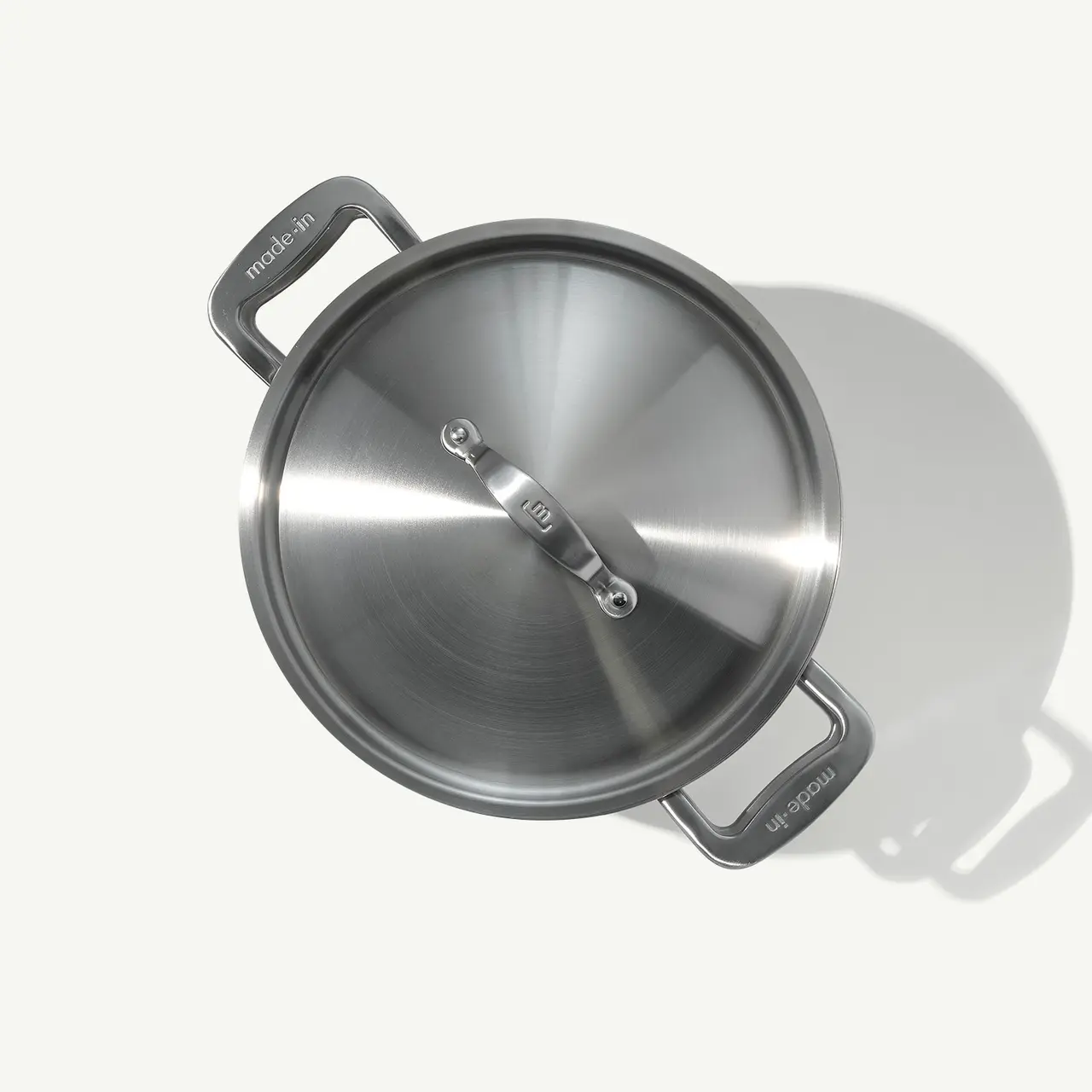 A stainless steel cooking pot with two handles sits on a light surface, casting a soft shadow.