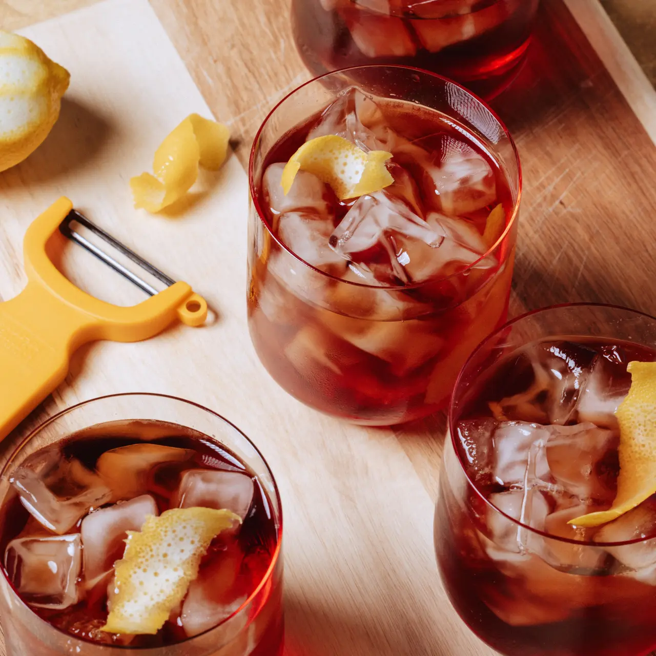 Five glasses of iced dark beverages garnished with lemon peel are arranged on a wooden surface, accompanied by a yellow peeler and a whole lemon.