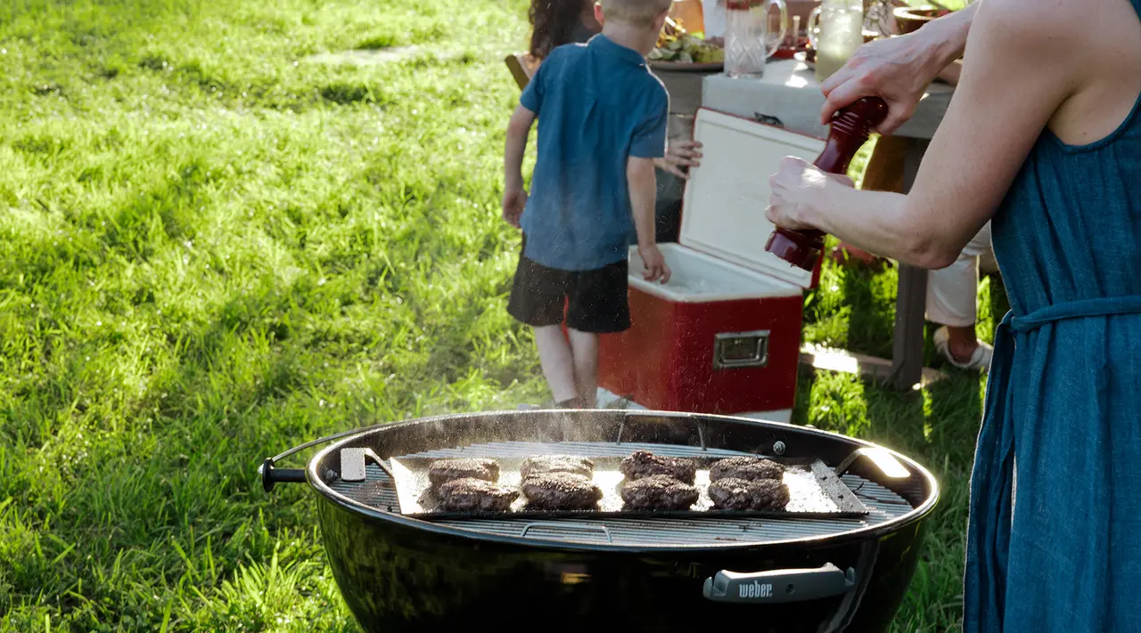 Burgers are grilling on an outdoor barbecue with a person seasoning them and a child in the background by a cooler on a sunny day.