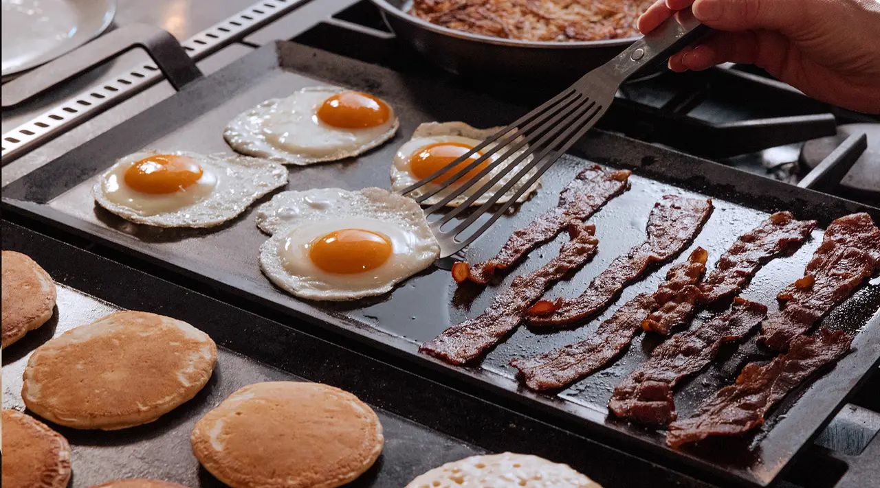 A variety of breakfast foods including pancakes, eggs, and bacon are being cooked on a large griddle, with a person's hand flipping an egg using a spatula.