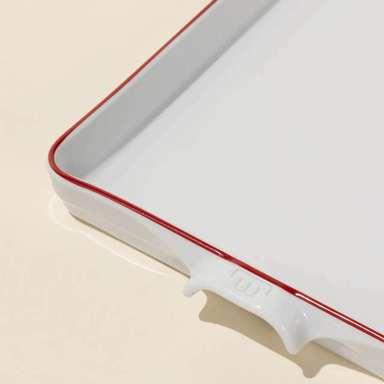 A white ceramic baking dish with red trim rests on a beige surface, angled to show the brand imprint on one handle.