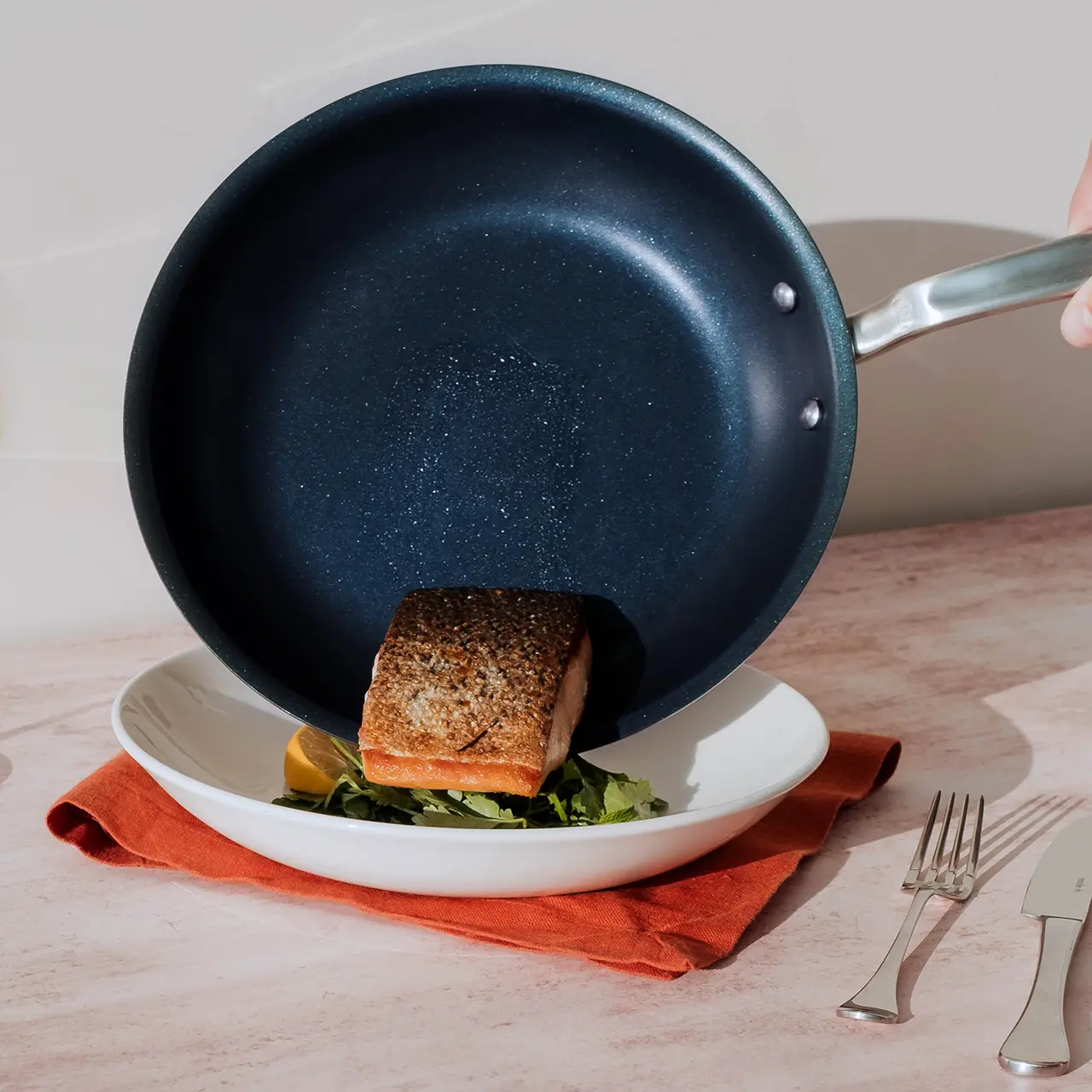 A seared piece of fish rests atop a bed of greens on a white plate, partially covered by a tilted blue frying pan, with a fork and knife placed beside on a light surface with a red napkin underneath.