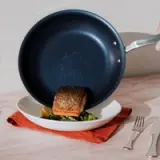 A seared piece of fish rests atop a bed of greens on a white plate, partially covered by a tilted blue frying pan, with a fork and knife placed beside on a light surface with a red napkin underneath.