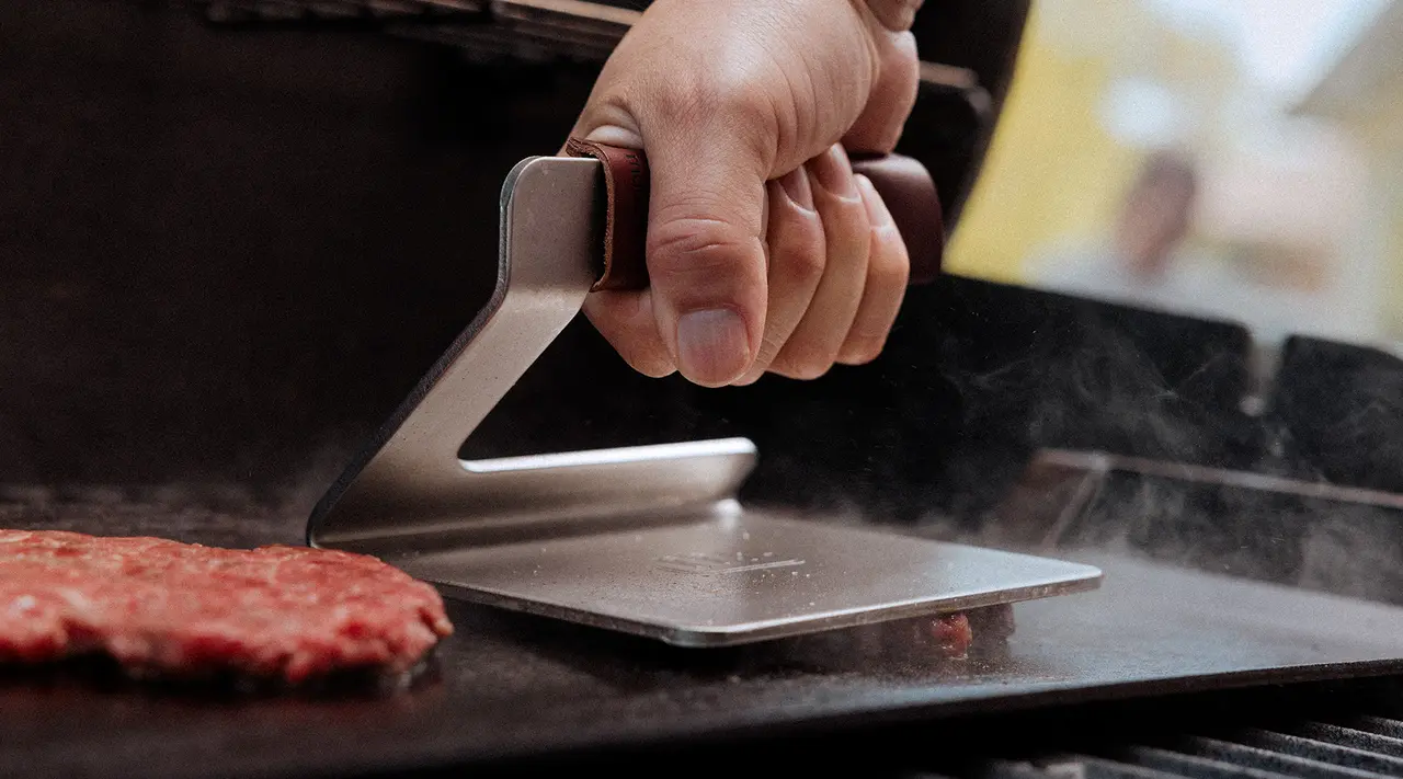 A person's hand presses a metal burger press onto a patty on a grill, causing steam to rise.