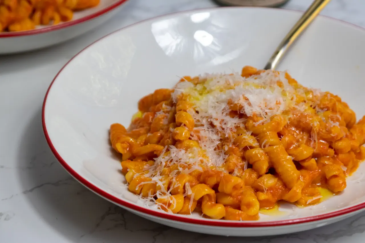 A plate of pasta with tomato sauce topped with grated cheese, with another similar plate in the background, both on a marble surface.