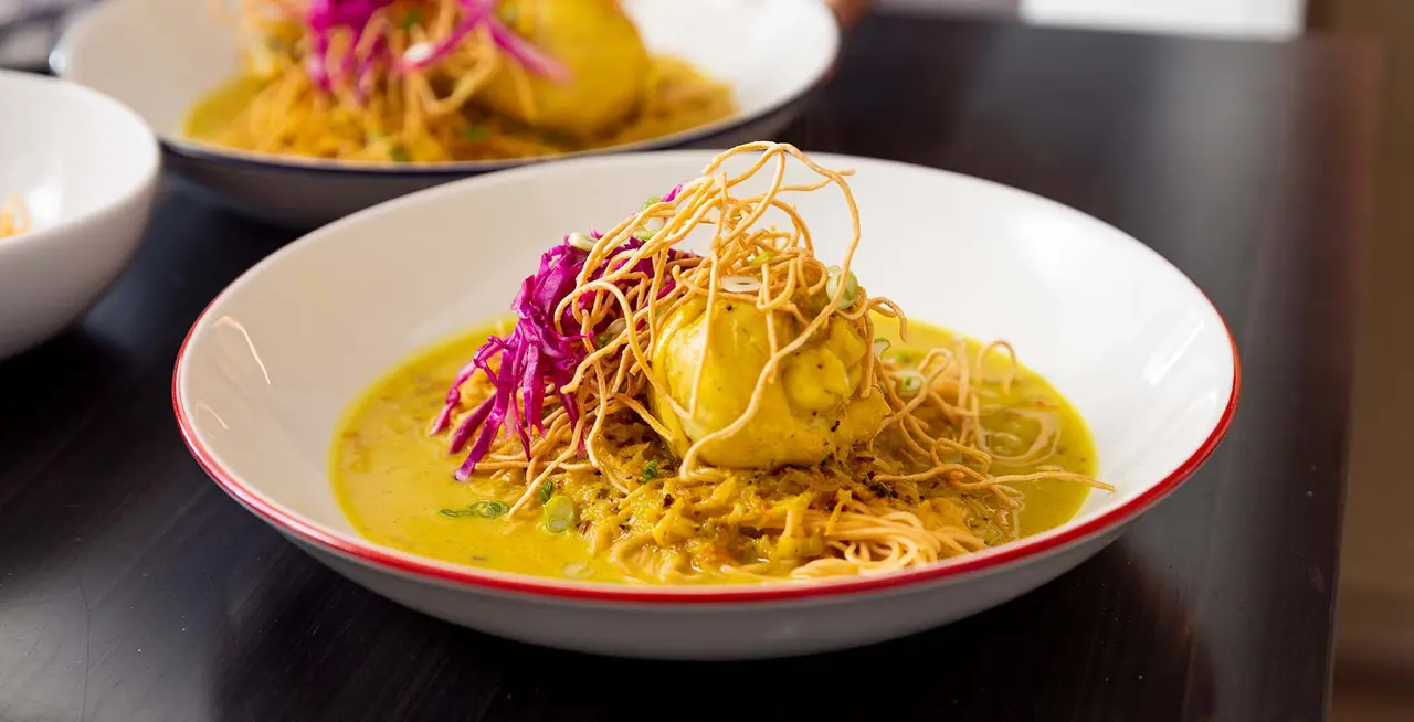 A colorful plate of food, possibly a curry, is garnished with purple slaw and crispy noodles, presented on a dark wood table.