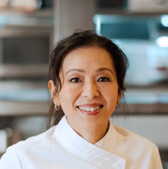 A smiling woman wearing a chef's uniform stands in a kitchen environment.