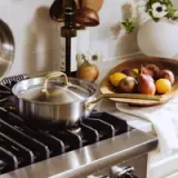 A stainless steel pot with a lid sits on a gas stovetop next to a wooden bowl filled with colorful fruits and a bottle of olive oil.