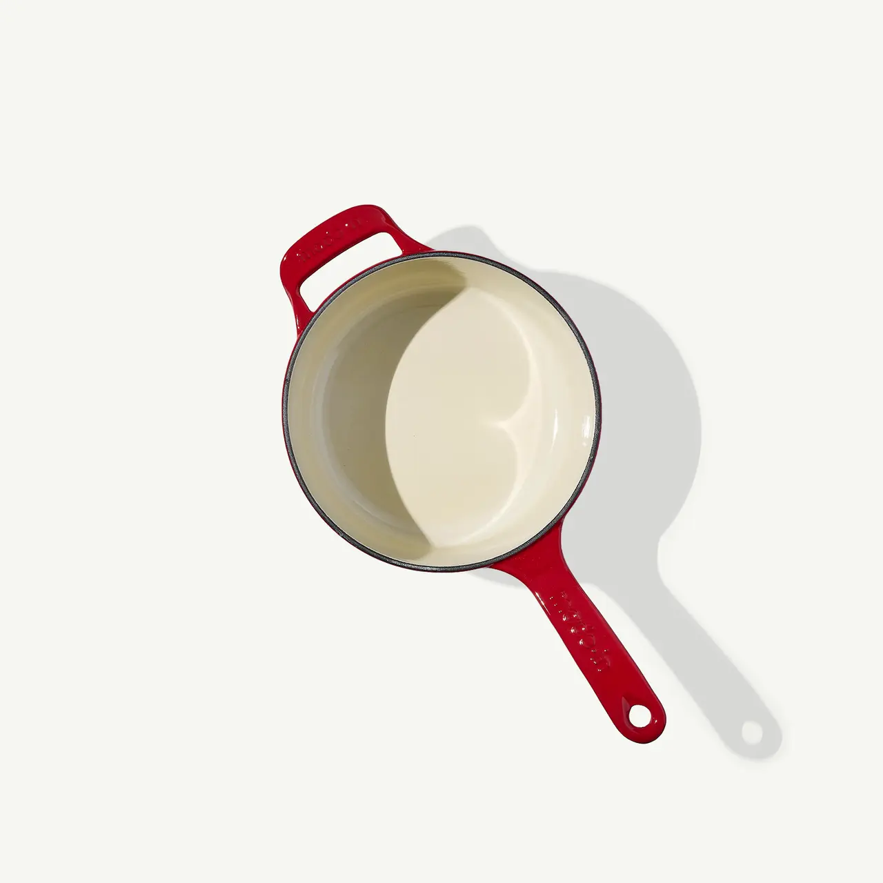 A red-handled saucepan is sitting on a plain surface casting a slight shadow, viewed from above.
