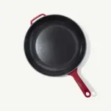 A round cast iron skillet with a red handle and a smooth black cooking surface rests on a light background.