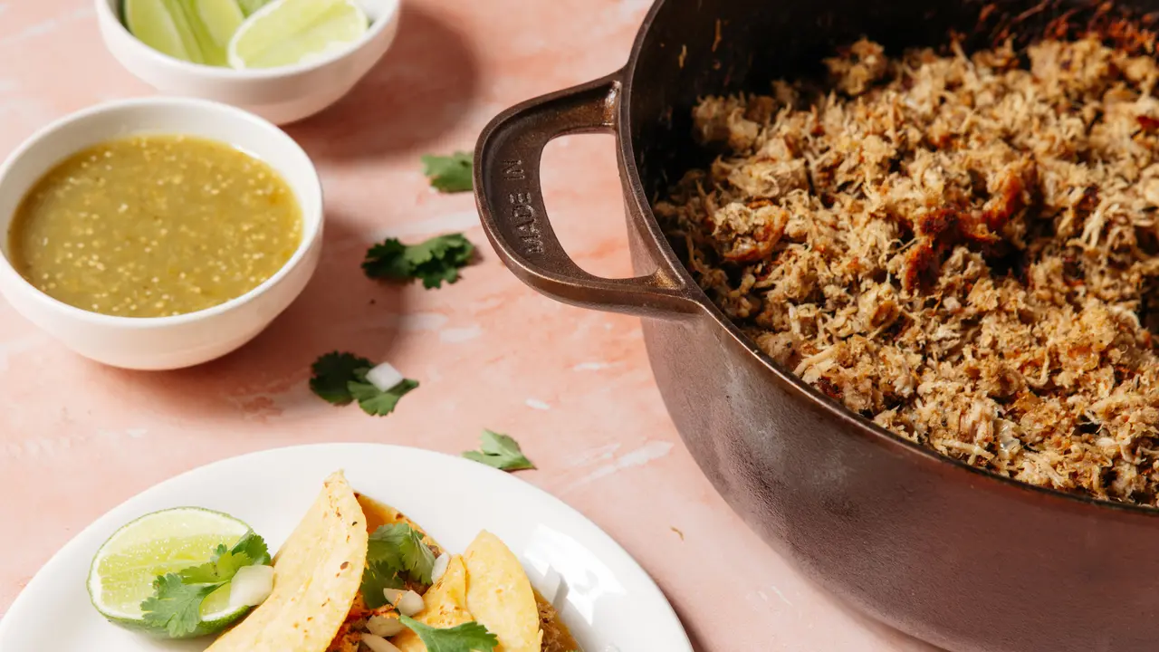 Made In Dutch Oven filled with shredded pork carnitas, beside salsa, lime, cilantro, and tortillas.