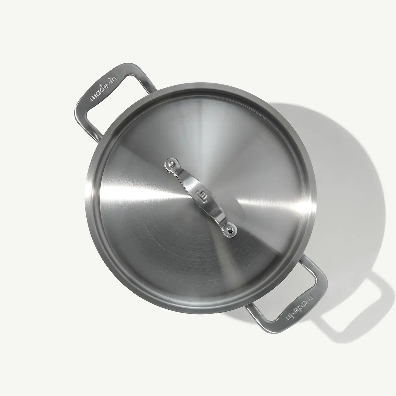 A stainless steel pot with handles and a lid casting a shadow on a light surface.