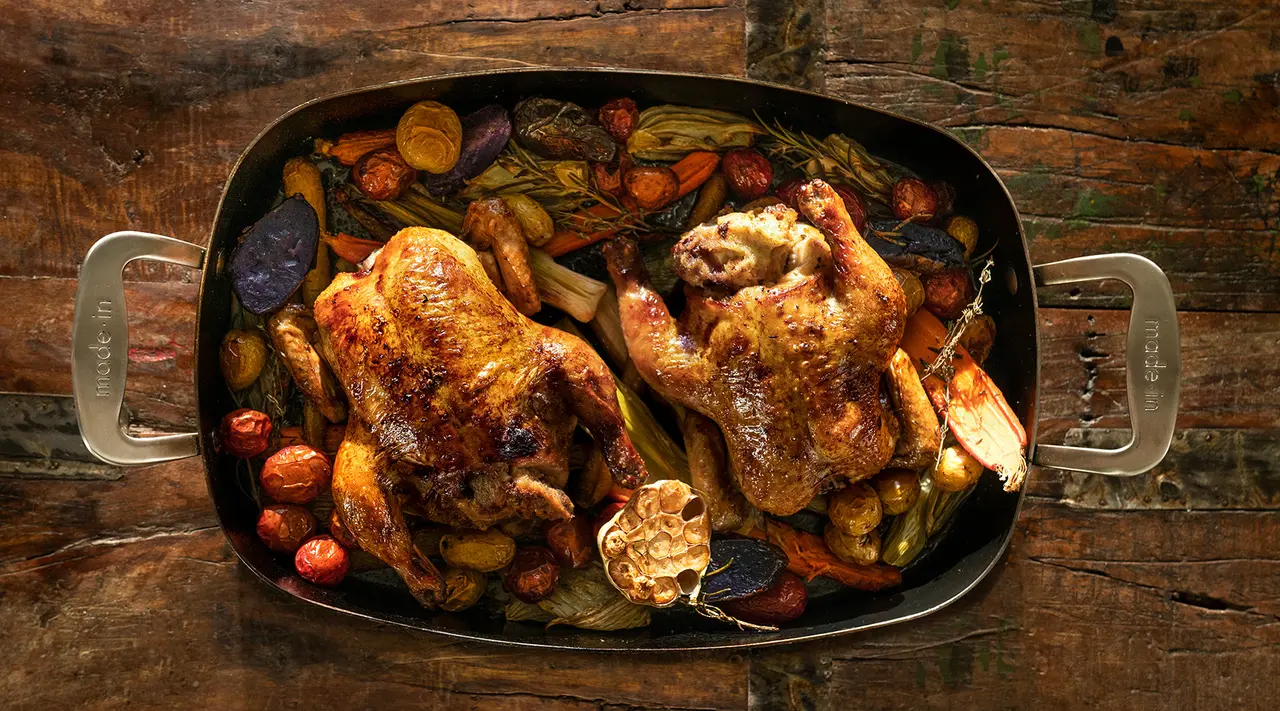 Roast chickens with a colorful assortment of roasted vegetables presented in a large oval roasting pan on a rustic wooden background.