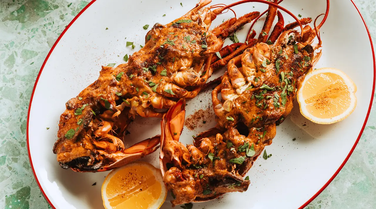 Two spiced, grilled lobsters are served on a platter with lemon wedges and garnished with herbs.