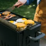A person is using a spatula to cook burgers, eggs, and bacon on a grill outdoors.
