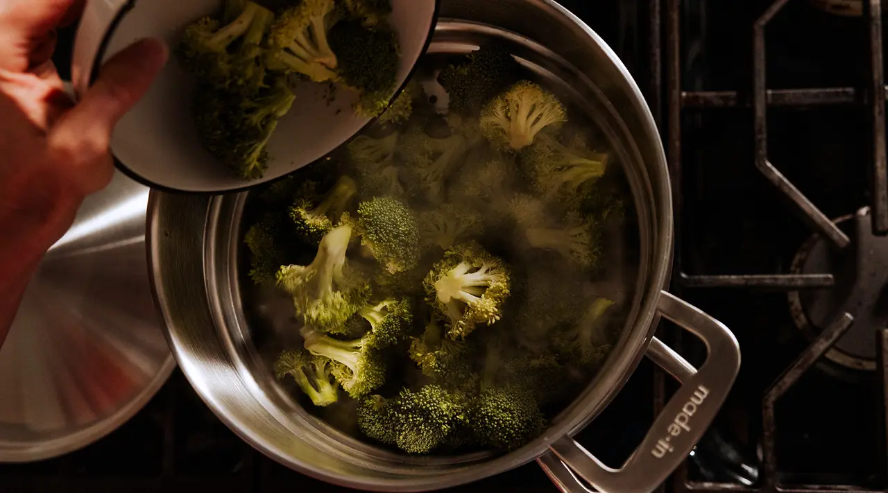 A person is adding broccoli florets to a pot of boiling water on a stove.