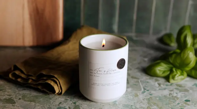 A lit scented candle sits on a marbled surface next to fresh basil leaves and a folded cloth.