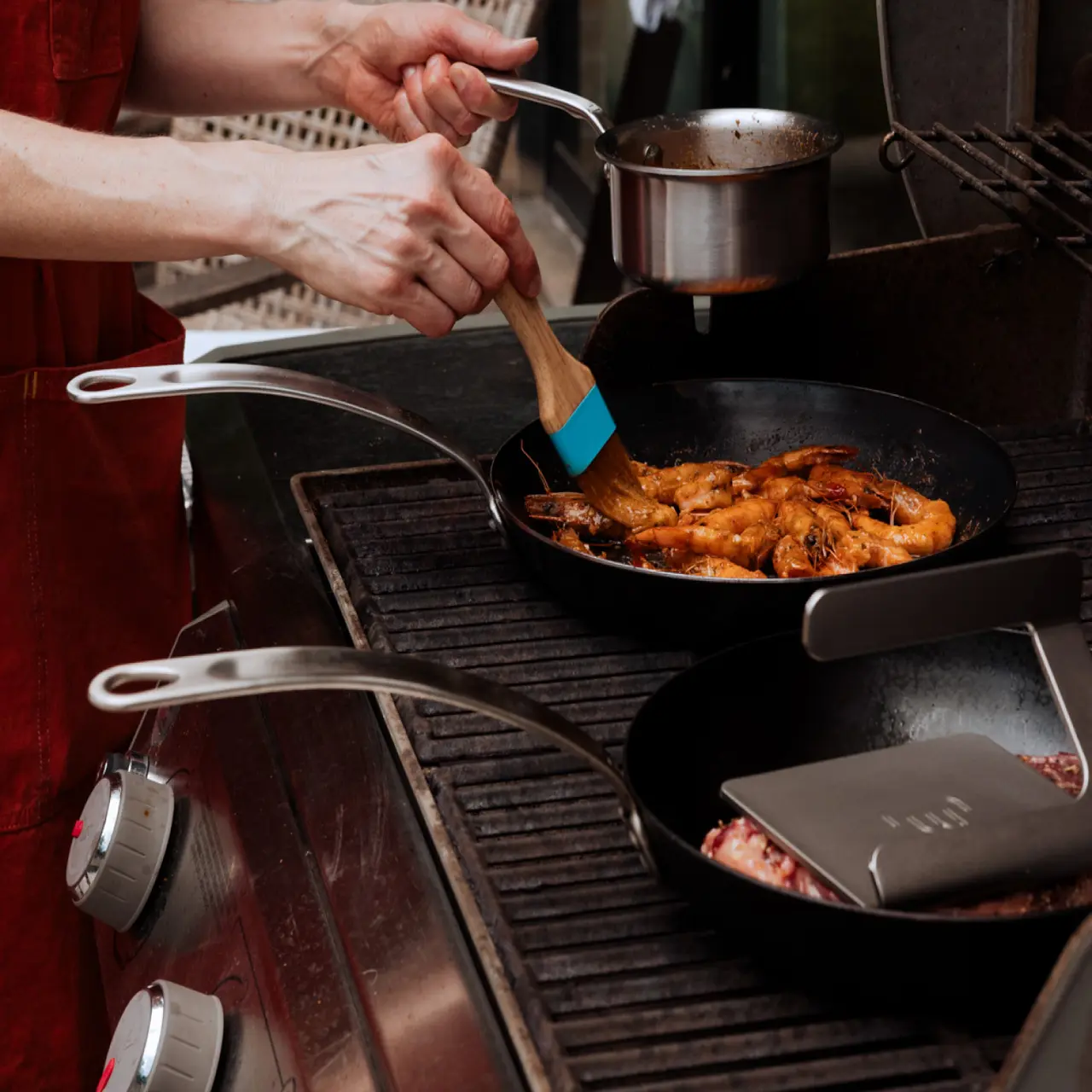 A person in a red apron is stirring shrimp in a skillet on a stove while other pots and pans are being heated nearby.