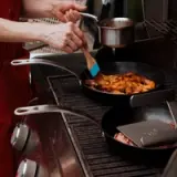 A person in a red apron is stirring shrimp in a skillet on a stove while other pots and pans are being heated nearby.