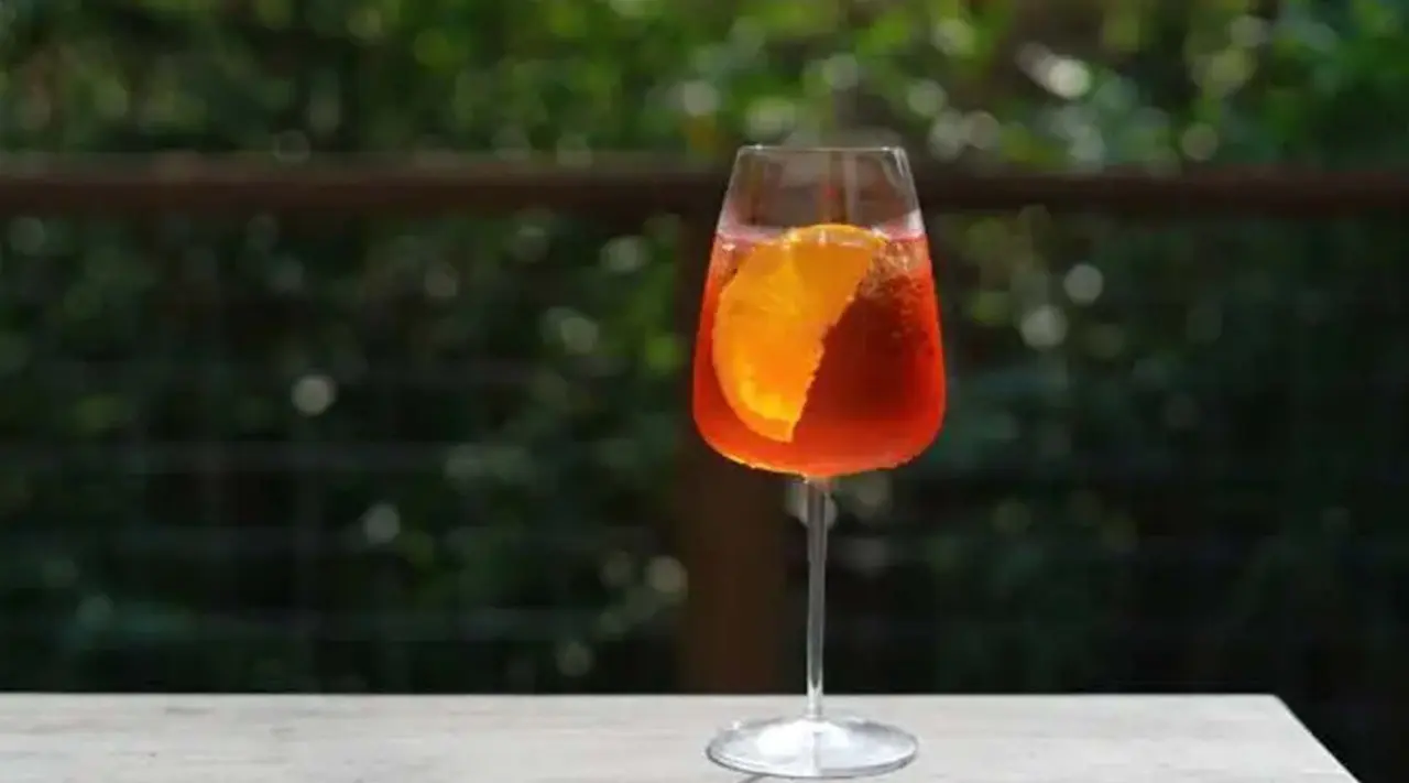 A glass of Aperol spritz with an orange slice sits on a wooden surface against a blurred greenery background.