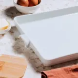 A baking scene with an empty white ceramic dish, scattered flour, eggs, a shell, butter, and a wooden spatula on a kitchen counter.