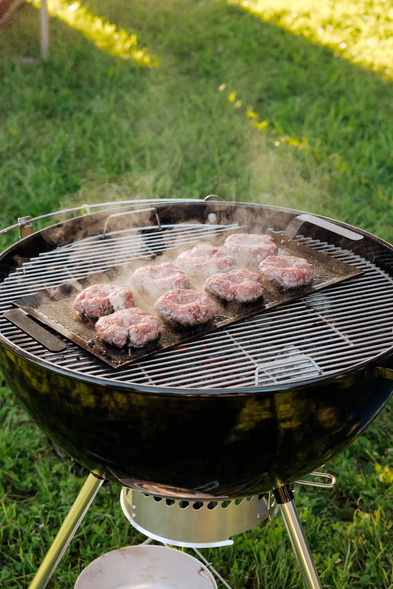 Burgers sizzle on a charcoal grill outdoors, with smoke rising against a green grass background.