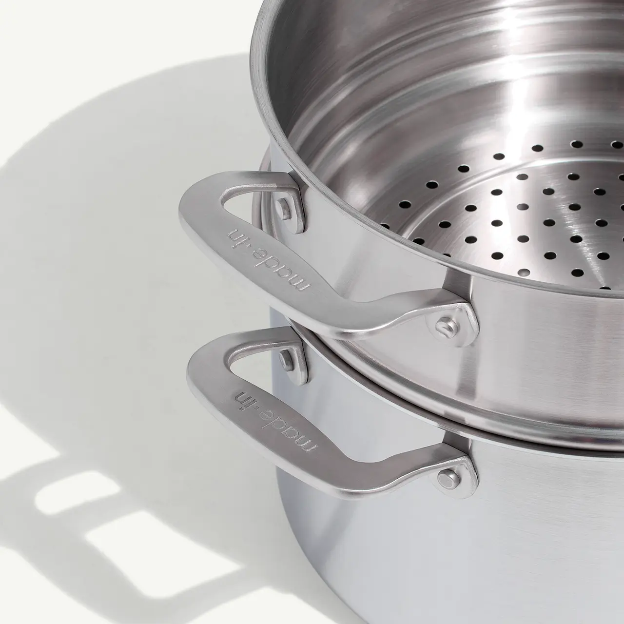 A stainless steel steamer pot with perforations and sturdy handles sits on a white surface, casting a shadow to its right.