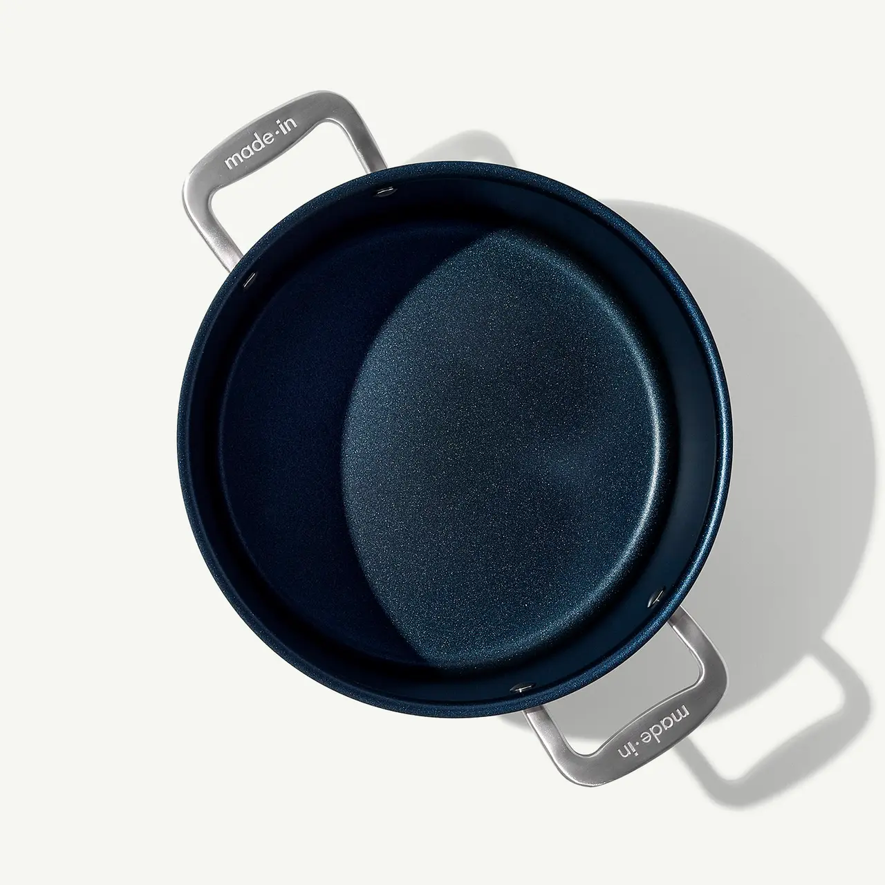 A non-stick frying pan is shown from above, casting a shadow on a light surface, with visible handles and brand markings.