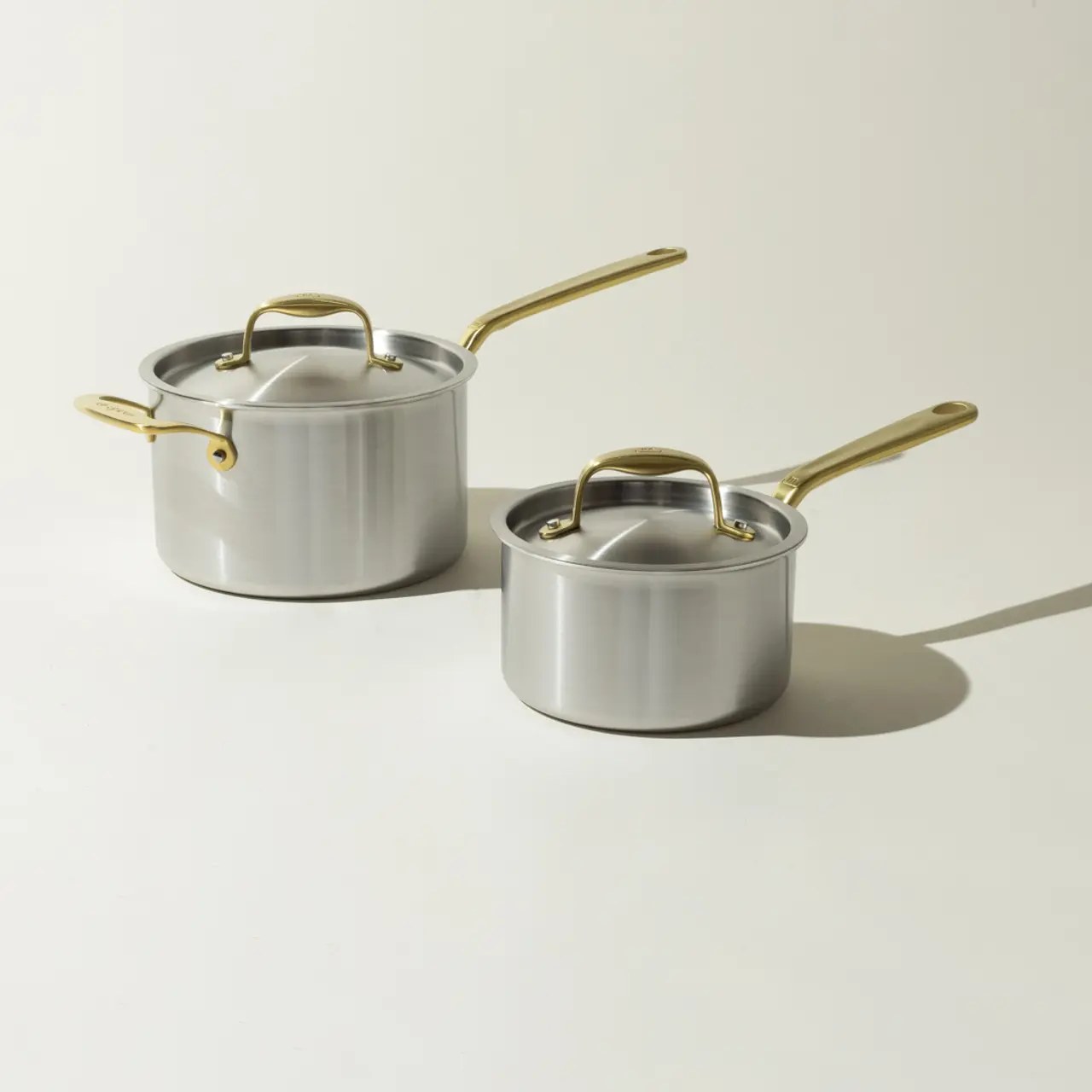 Two silver saucepans with golden handles cast soft shadows on a light background.