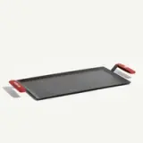 A rectangular cooking griddle with red handles on either side lies flat on a neutral background.