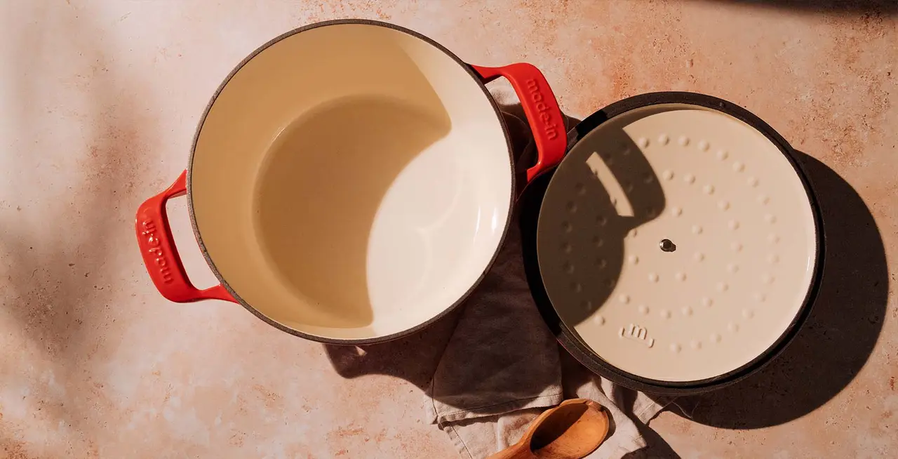A cream-colored Dutch oven with red handles is placed beside its inverted black lid, resting on a beige surface, with a wooden utensil nearby.