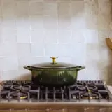A green, lidded cooking pot sits on a modern gas stove against a backdrop of light-colored tiled walls.