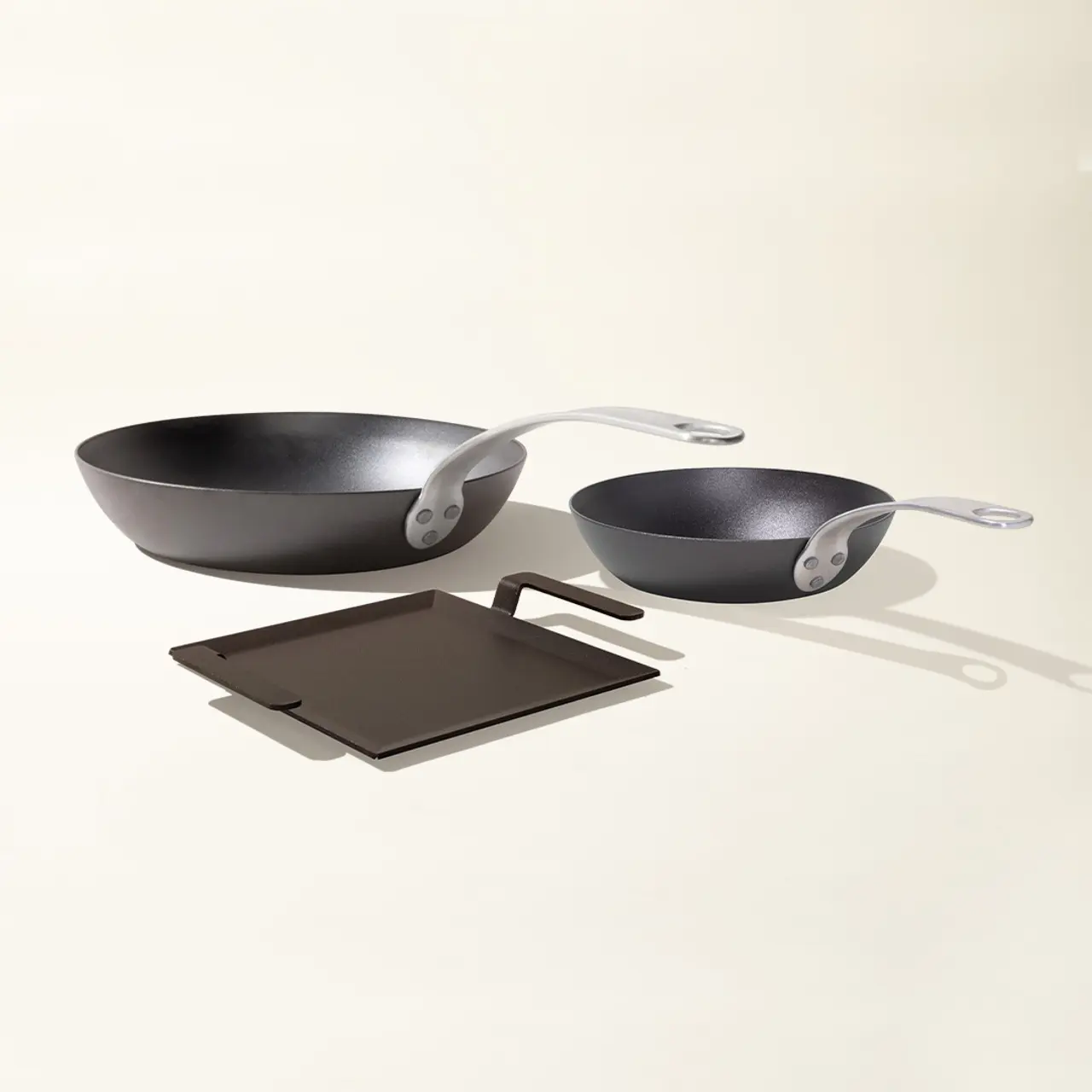 Two different-sized, black frying pans with silver handles rest on a light surface alongside a dark square trivet.