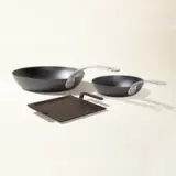 Two different-sized, black frying pans with silver handles rest on a light surface alongside a dark square trivet.