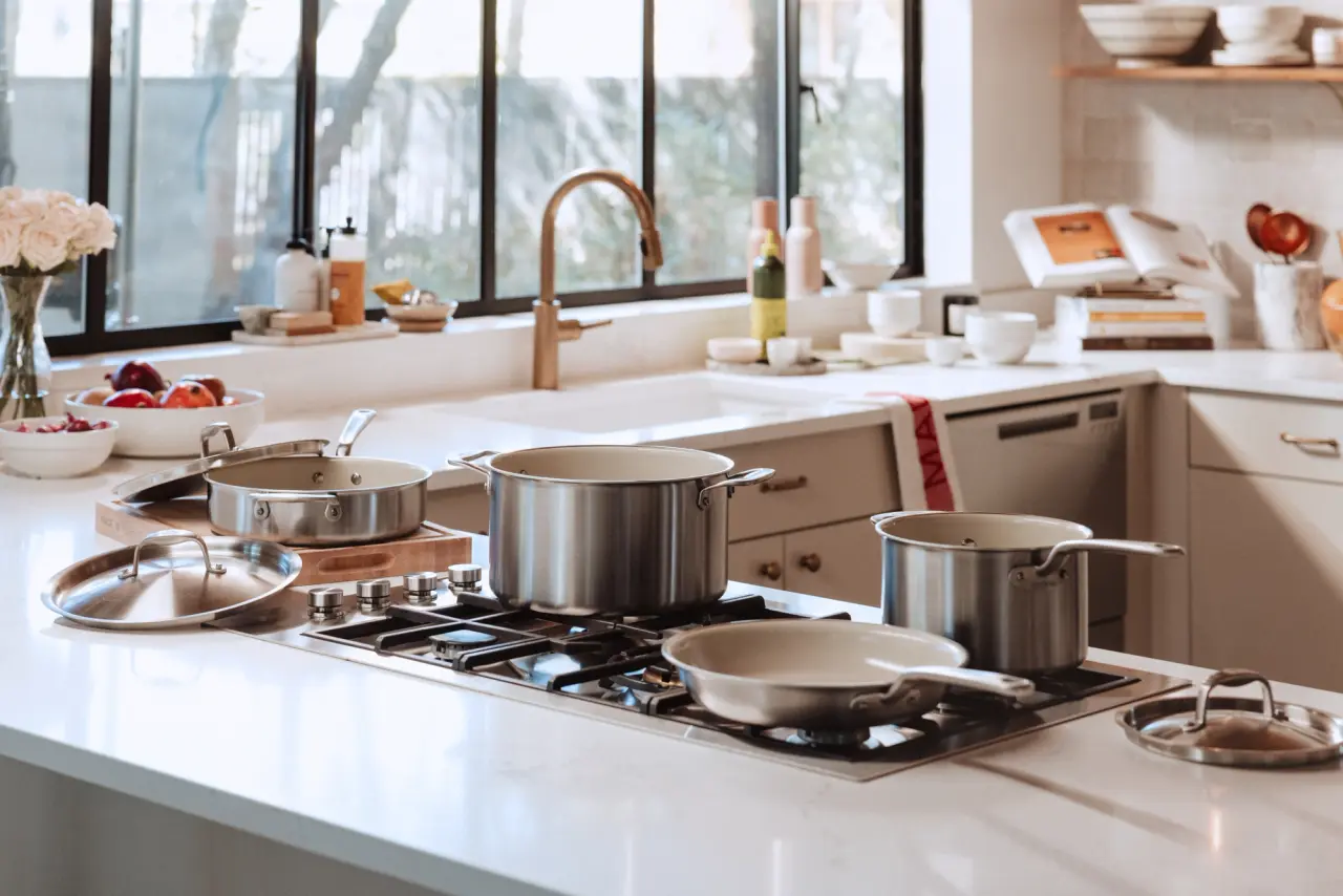 A modern kitchen counter features various stainless steel pots and pans on a stove, surrounded by fresh produce and kitchenware.