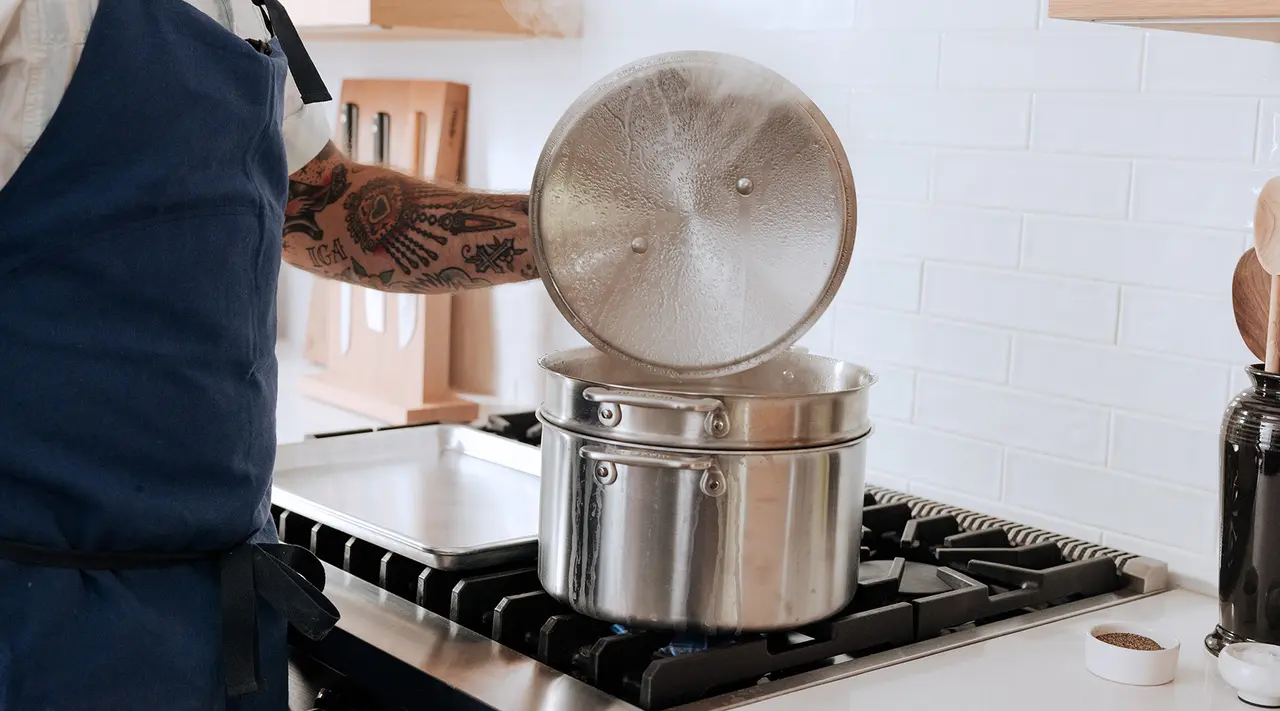 A tattooed arm lifts the lid of a steaming pot on a gas stove in a home kitchen.