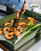 A person is using tongs to lift cooked shrimp and green onions from a grill tray.